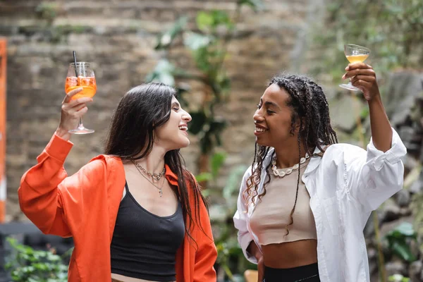 Smiling interracial girlfriends holding cocktails and looking at each other on cafe terrace — Foto stock