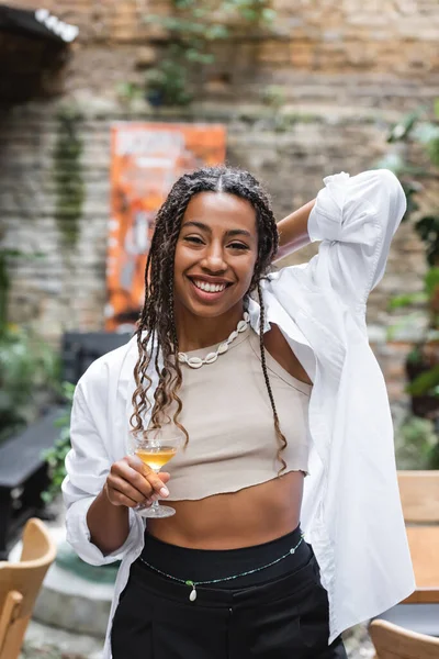 Cheerful african american woman holding cocktail on terrace of cafe - foto de stock