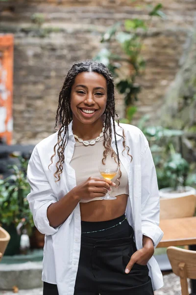 Positive african american woman holding glass of cocktail on terrace of cafe — Photo de stock