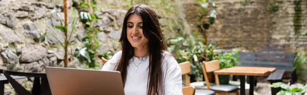 Cheerful brunette freelancer using laptop on terrace of cafe, banner — Photo de stock