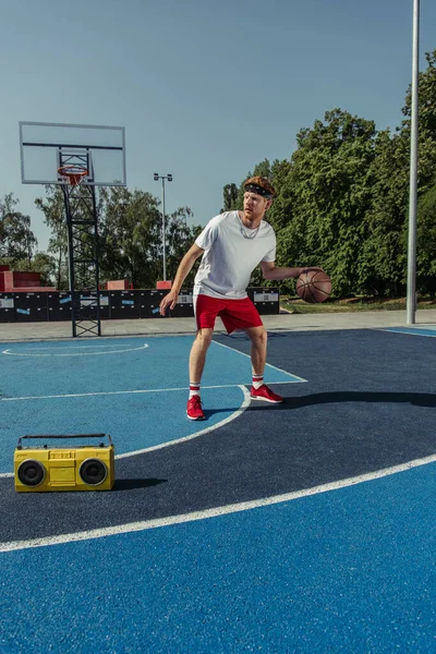 Young sportsman playing basketball near tape recorder on modern court — Stockfoto