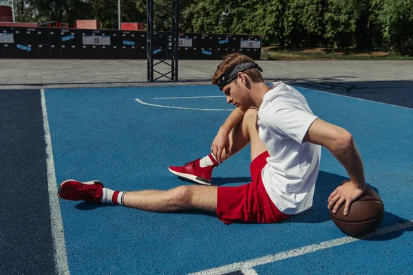 Баскетболист в красных шортах и кроссовках сидит на площадке рядом с мячом — стоковое фото