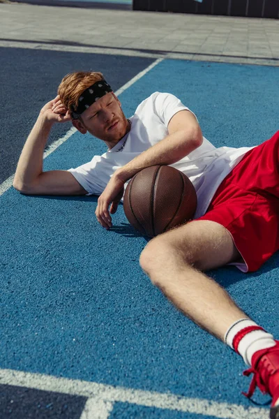 Redhead basketball player in sportswear lying on court near ball and looking away — Foto stock