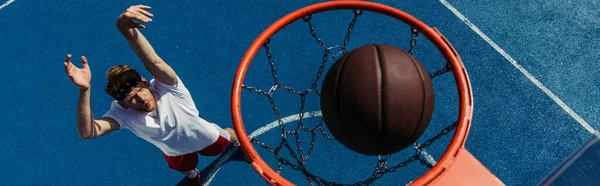 Top view of ball in basketball hoop above young man training on court, banner — Photo de stock