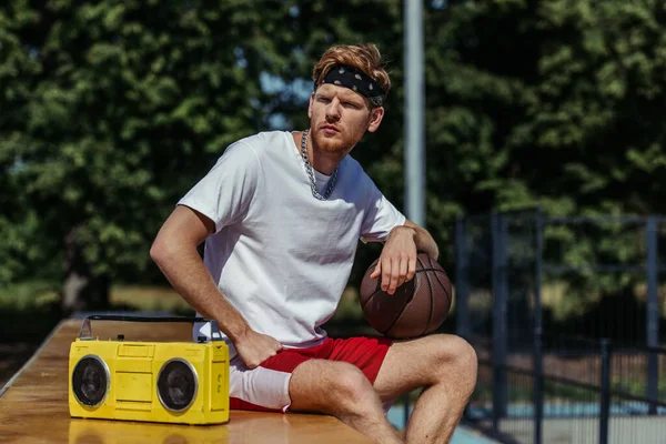 Young basketball player sitting with ball near tape recorder and looking away - foto de stock