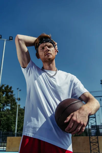 Low angle view of basketball player in white t-shirt and necklace holding ball outdoors — Stock Photo
