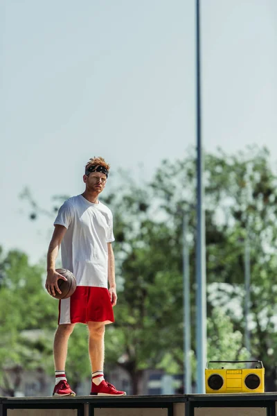 Повна довжина людини в спортивному одязі, що тримає м'яч, стоячи біля бумбоксу — стокове фото