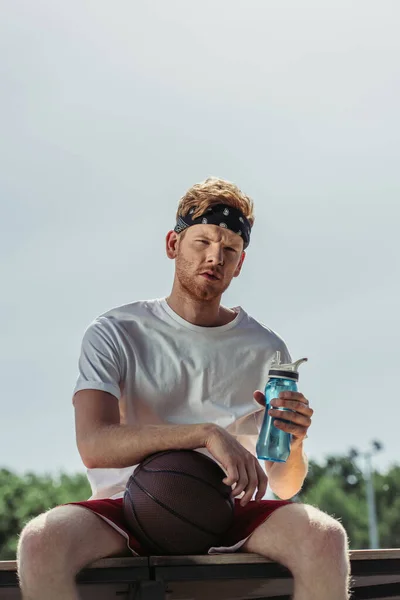 Sportive man in bandana holding sports bottle while sitting with ball — Fotografia de Stock