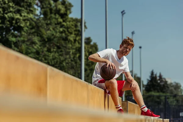 Young basketball player with ball sitting on stadium on blurred foreground — Stock Photo
