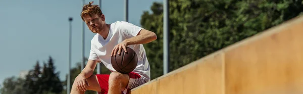 Рыжая баскетболистка, сидящая на стадионе с мячом, баннером — стоковое фото