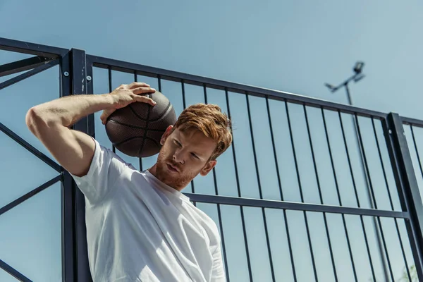 Low angle view of redhead man in white t-shirt standing with ball near fence — Foto stock