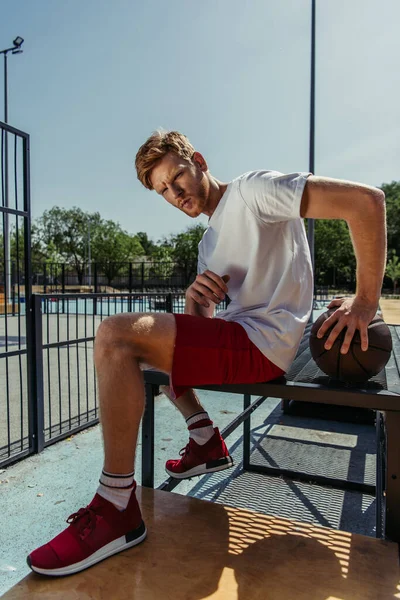 Full length of young redhead sportsman sitting on bench with ball — Foto stock