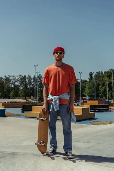Full length of trendy man in sunglasses standing with skateboard in skate park — Stock Photo