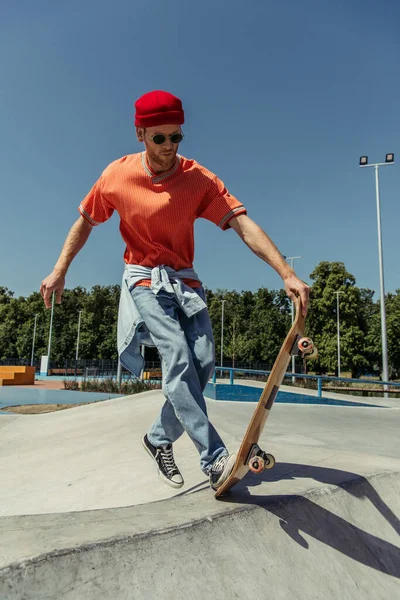 Полная длина человека в модной одежде и солнцезащитных очках тренировки со скейтом в парке — стоковое фото
