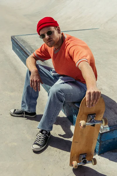 Man in sunglasses and red beanie sitting with skate and looking at camera — Foto stock