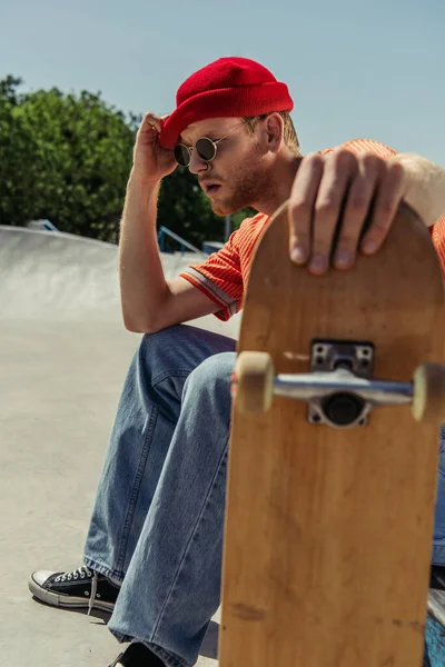 Exhausted man sitting with blurred skateboard and touching beanie — Stock Photo