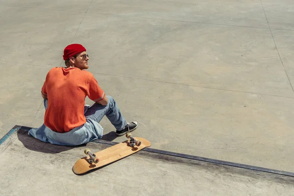 Back view of man in orange t-shirt and red beanie sitting near skateboard on ramp — Stock Photo