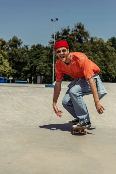 Excited man in red beanie and sunglasses riding skateboard in skate park — Stock Photo