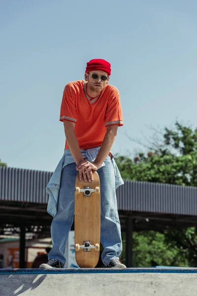 Full length of man in trendy clothes and sunglasses standing with skate in park — Stock Photo