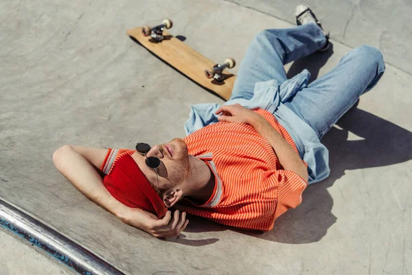 High angle view of trendy man in sunglasses relaxing on ramp near skateboard — Stockfoto