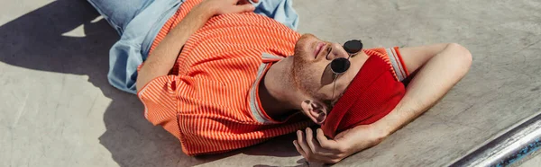 Trendy skateboarder in red beanie and sunglasses lying on ramp in skate park, banner — Fotografia de Stock