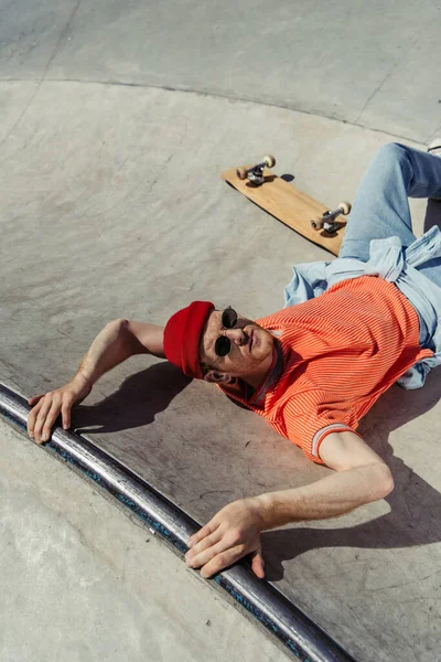 High angle view of man in sunglasses and beanie looking at camera while lying on ramp near skate — Stockfoto