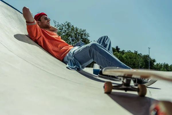 Low angle view of happy and stylish man lying on ramp near blurred skateboard — Fotografia de Stock