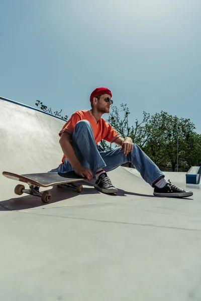 Full length of trendy man in gumshoes and sunglasses sitting near skateboard - foto de stock