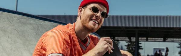 Trendy skateboarder in red beanie and sunglasses smiling outdoors, banner — Stock Photo
