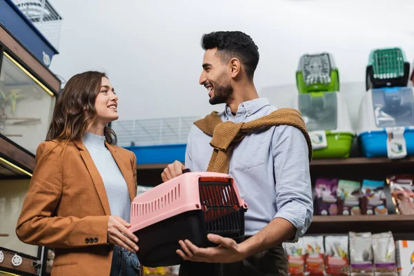 Cheerful interracial couple talking while holding animal cage in pet shop — Stock Photo
