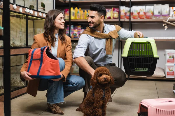 Smiling multiethnic couple holding bag and cage near poodle in pet shop — Stock Photo