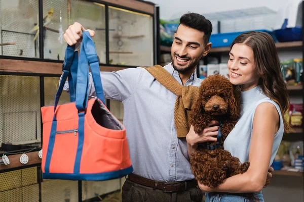 Smiling woman holding poodle near muslim boyfriend with animal bag in store — Stock Photo
