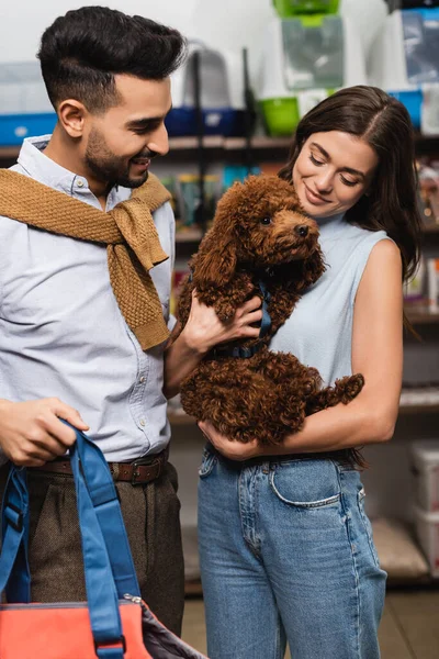 Улыбающаяся женщина держит пуделя рядом с мусульманским парнем с сумкой в магазине животных — стоковое фото