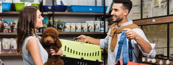 Side view of cheerful interracial couple holding poodle and animal cage in store, banner — Stock Photo