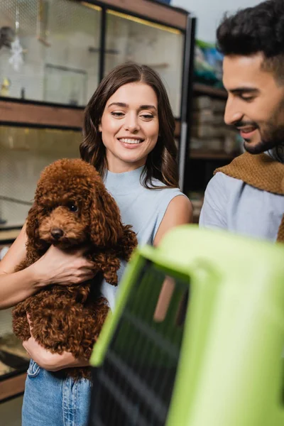 Smiling woman holding poodle near arabian boyfriend and cage in animal shop — Stockfoto