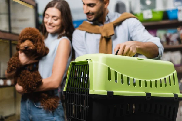 Blurred multiethnic couple with poodle holding animal cage in shop — Stockfoto