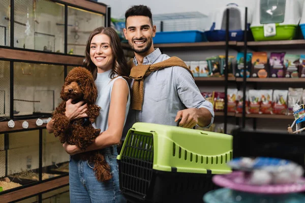 Positive interracial couple holding animal cage and poodle in pet shop — Stock Photo