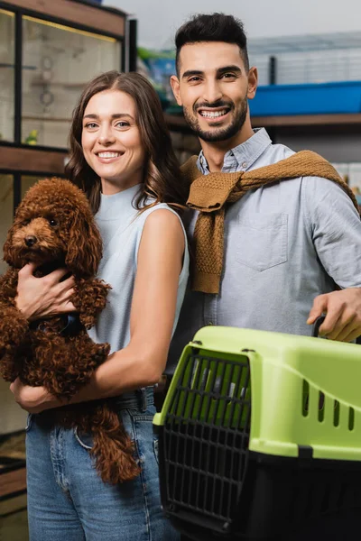 Cheerful multiethnic couple holding poodle and animal cage in pet shop — Stock Photo
