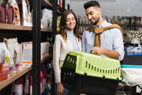 Smiling multiethnic couple looking at animal cage near showcase in pet shop — Stock Photo