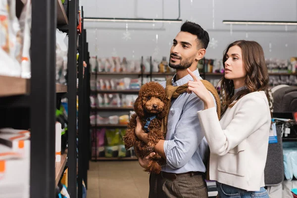 Woman pointing with finger near muslim boyfriend holding poodle in pet shop — Stockfoto