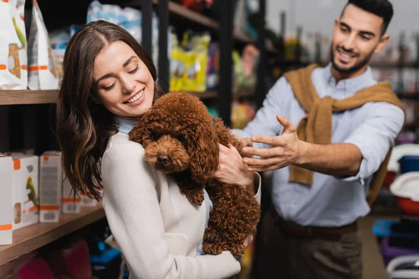 Smiling woman holding poodle near blurred arabian boyfriend and packages in pet shop - foto de stock