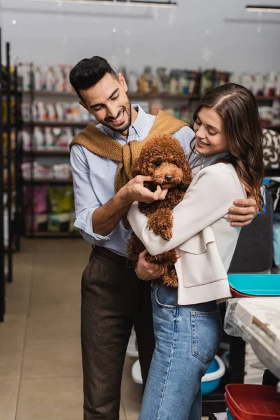 Cheerful multiethnic couple holding poodle in pet shop — Stockfoto