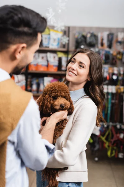 Улыбающаяся женщина держит пуделя рядом с расплывчатым парнем в магазине животных — стоковое фото