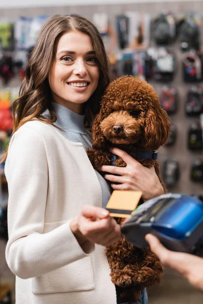 Cheerful woman holding poodle and credit card near payment terminal in pet shop - foto de stock