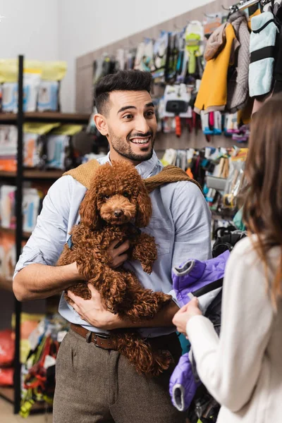 Excited muslim man holding poodle near blurred girlfriend with animal jacket in pet shop — Photo de stock