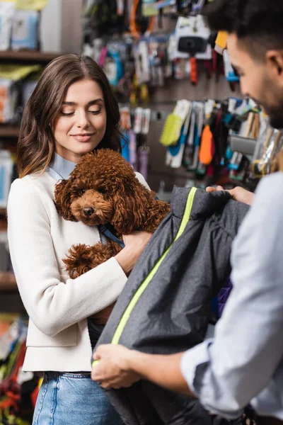 Smiling woman holding poodle near arabian boyfriend with animal jacket in pet shop — Foto stock