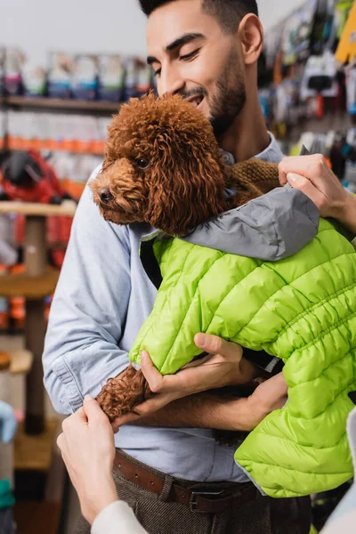 Woman wearing animal jacket on poodle near arabian boyfriend in pet shop — Foto stock