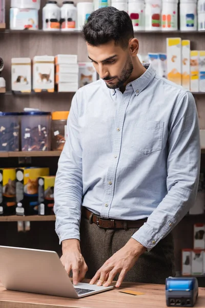 Arabian seller using laptop near payment terminal in pet shop — Stockfoto