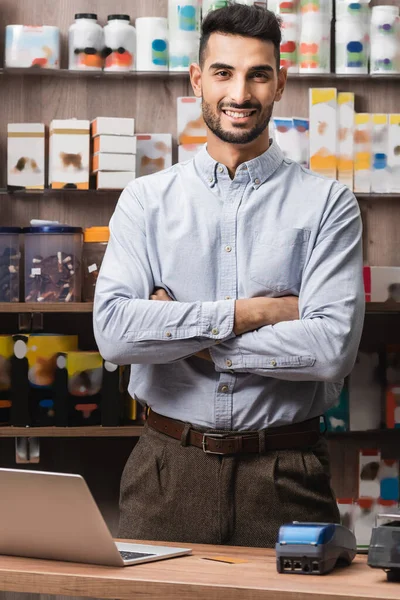 Smiling muslim seller crossing arms near laptop in pet shop - foto de stock