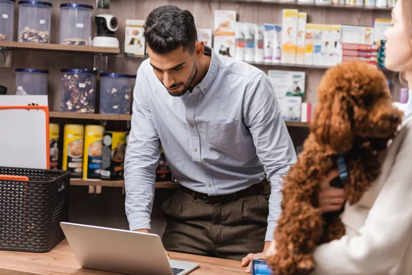 Arabian salesman looking at laptop near blurred customer with poodle in pet shop - foto de stock